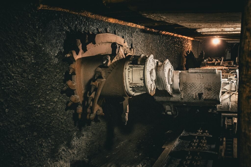 Close-up of a coal cutting machine's toothed drum with rusted steel picks, positioned against a coal seam wall in a dimly lit underground mine tunnel, with warm lighting illuminating the passage in the background.