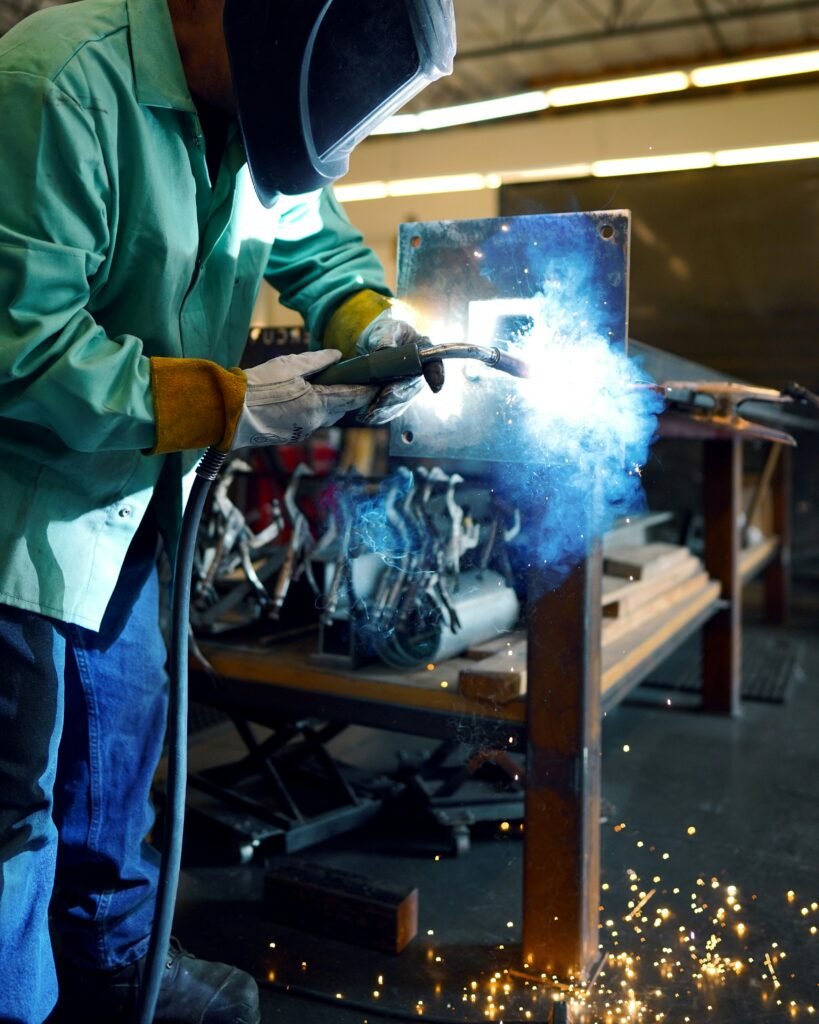 `Industrial engineer performing metal welding in a manufacturing workshop while wearing protective gloves, clothing, and a safety helmet.`